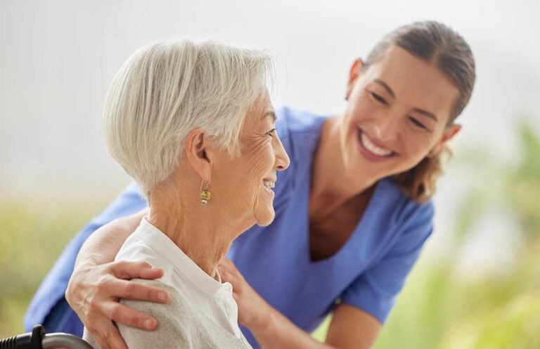 Older adult speaking with a caregiver in a bright assisted living common room beside a dining table and walker